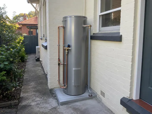 Outdoor cylindrical hot water tank on a concrete pad beside a cream brick house in Five Dock